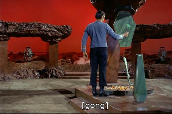 Star Trek The Original Series scene. We're on a planet that appears rocky with a red backdrop sky because it was probably inexpensive! We're looking at some kind of ceremonial area, with stone carved features and flat surface for a performance or ritual. In the middle is maybe a fire or coal pit, and some artifacty items. Standing with his back to us is Spock, in baby blue uniform, and he's hitting a mallet against one of those gong things that makes a big gong sound. Closed caption reads, "[gong]"