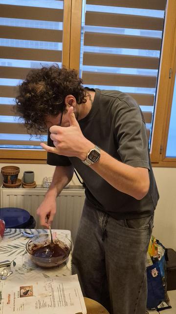 Un jeune homme en train de battre une pâte à gâteau au chocolat "maison" faisant 1 signe de la main pour signifier qu'il n'est pas super d'accord pour qu'on le prenne en photo.