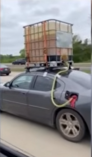 It's a dark metallic grey car with a large industrial tank made out of plastic with an aluminum grid strengthening the plastic mounted to the top of the car filled with a a yellowish liquid that's probably gasoline sloshing around in the tank. A yellow hose leads from the tank and into the cars fuel tank.