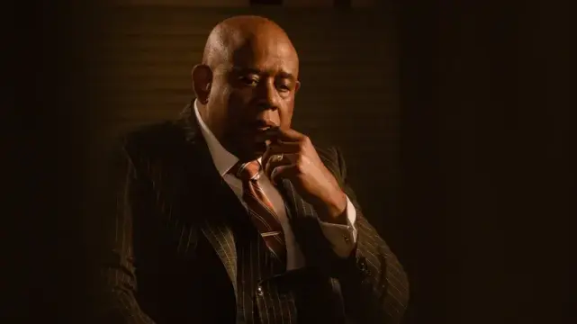 Forest Whitaker as 'Bumpy' Johnson in "Godfather of Harlem", wearing a pinstripe suit and maroon tie, sitting in moody low light, fingers near his lips, contemplative.
