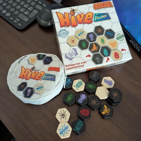 La imagen muestra el juego de mesa "Hive Pocket Ultimate" desplegado sobre una mesa de madera. En la parte superior derecha se encuentra la caja del juego, blanca con el logo "Hive" grande en naranja y "Pocket" en azul, rodeado de un patrón hexagonal y ejemplos de las fichas de insectos. La caja indica que incluye las tres expansiones y una bolsa de viaje. A la izquierda, hay una pequeña bolsa de tela blanca, redonda, con el mismo logo y algunas fichas de insectos impresas. En la parte inferior de la imagen, numerosas fichas hexagonales del juego están dispersas. Estas fichas son de dos colores principales, crema claro y negro, y cada una tiene la ilustración de un insecto diferente en un color vibrante, como arañas, hormigas, saltamontes, mariquitas y abejas.
