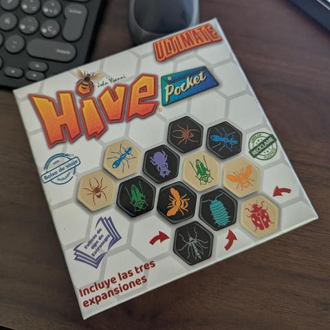 Una caja del juego de mesa "Hive Pocket Ultimate" se muestra ligeramente inclinada sobre un escritorio de madera oscura. La caja es predominantemente blanca con un patrón hexagonal de fondo. El logo principal "Hive" es grande, naranja y tiene textura de panal, con la palabra "Ultimate" en rojo arriba y "Pocket" en azul a la derecha. Una abeja vuela cerca del logo. La caja exhibe varias fichas hexagonales de juego, algunas de color crema y otras negras, cada una con una ilustración de un insecto diferente como arañas, hormigas, escarabajos, abejas, mariquitas y más. El texto en español en la caja destaca: "Incluye las tres expansiones", "con Bolsa de viaje incluido", y "Folleto de tips de Estrategia". También hay un sello verde que dice "Libre de PVC Reciclable Plástico". Parte de un teclado de computadora negro es visible en la esquina superior izquierda.