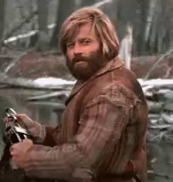 Robert Reford, in his iconic role as Jeremiah Johnson, sitting beside a pond, sporting a beard and mountain man's clothing. He nods and smile approvingly.