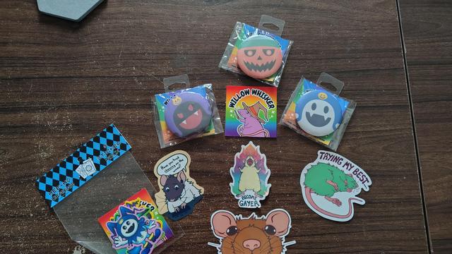 Pin badges and stickers of the Jack trio demons from SMT/Persona. Four stickers of rats, one is sprawled with text "Trying my best...". One is powering-up and yelling "Become Gayer!" and one is talking on a headset while typing, performing tech support with a curious expression saying, "Have you tried *chewing* on the wires?". Final rat sticker is a close-up front-on view of a rat peeking its head over whatever it gets stuck to.
