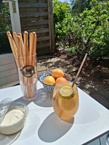 Coctail glass filled with Slices of Lime and Lemon with Tschunk mix inside of it. This is sitting on a table outside with a few snacks and dip on the table