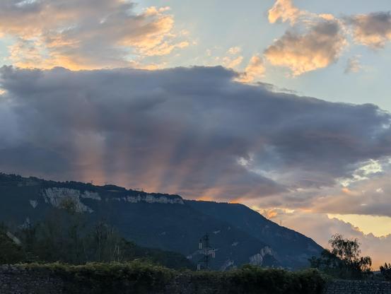 Soleil couchant sur le Vercors. En premier plan, la montagne. Un gros nuage gris au dessus.
Et entre les nuages et le profil des montagnes, des rayons lumineux orangés qui semblent sortir de la montagne tels des projecteurs.