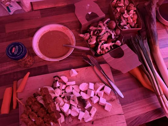 Eine Holzarbeitsplatte ist mit verschiedenen Lebensmitteln ausgestellt: eine Schüssel Suppe, ein Glas Pesto, gehackte Pilze, etwas gewürfeltes Gemüse, Fleischspieße und ganze Karotten und Lauch. Das Bild hat einen rosa Farbton.