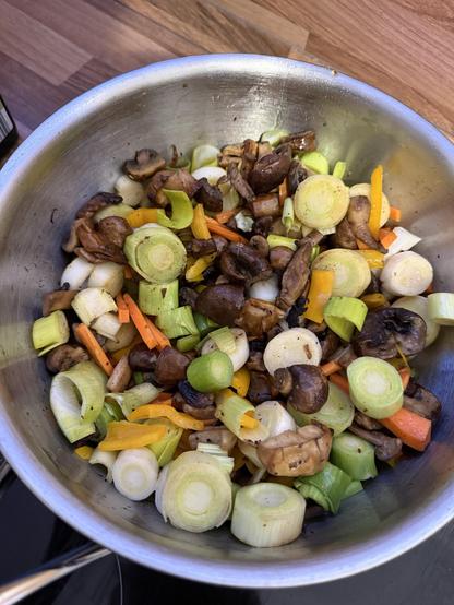 Eine Edelstahlschale mit einer Mischung aus sautiertem Gemüse, darunter Pilze, Lauch, Karotten und Paprika, vor einem Holzhintergrund.