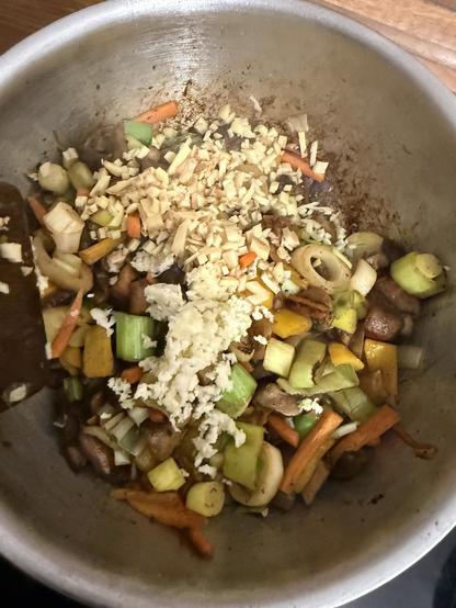 Ein Metalltopf gefüllt mit sautiertem Gemüse, darunter Karotten, Paprika und Sellerie, gekrönt mit gehacktem Ingwer und Knoblauch. Die Mischung scheint Teil eines Kochprozesses zu sein.