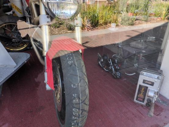 view through the window of a motorbike shop, showing the front tread and headlight of a racing bike; and down on the carpet next to it, a little toy replica of Cloud's bike from FF7 (the remake Hardy-Daytona, not Fenrir)