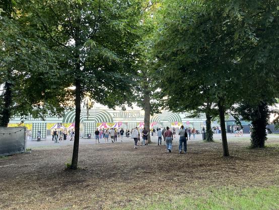 The festival entrace of Summerstage Groningen peaking through leafy trees, humans walking in front. The entrance sports a white and green colorway, with clean lines spread over the wooden front banners over the entrance lanes.