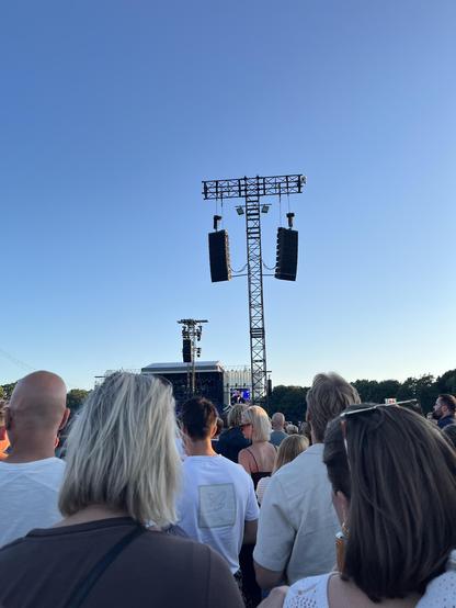 A huge metal T-shaped metal scaffold with two large sound speakers hanging from it. In the far back of the image is the stage with a huge display that shows a glimpse of singer Marcus Mumford. Loads of people inbetween.