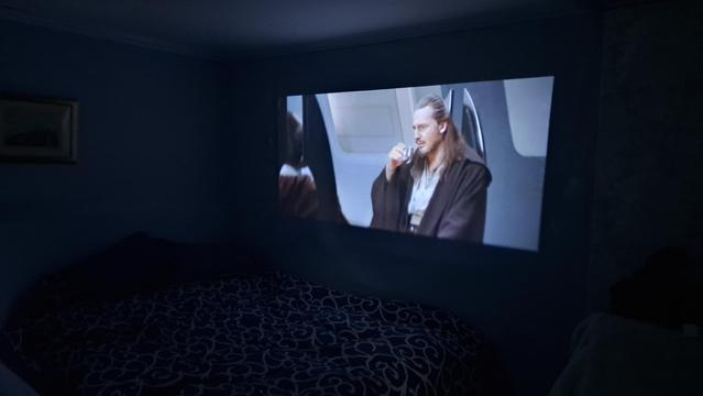 A dark room, but you can just make out a bed against the wall. And running the length of the bed, projected on to the wall, is a scene from "The Phantom Menace", where Qui Gon Jin sips at a drink.