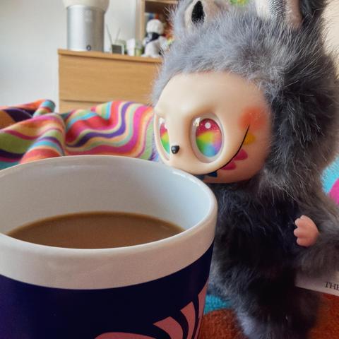 Eine bunte Decke ist im Hintergrund drapiert, mit einem lustigen Labubu mit regenbogenfarbenen Augen neben einer mit Kaffee gefüllten Tasse. Die Umgebung ist gemütlich und entspannt.
