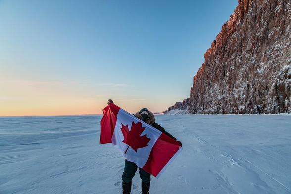 A man in a parka and toque, with a face mask, stands on a frozen ocean, and holds a Canadian flag. The sky is light blue and along the horizon is tinted with yellows, pinks, and oranges. Along the right side, 600 foot tall red cliffs stretch along to the horizon. (Photo taken in February for flag day)