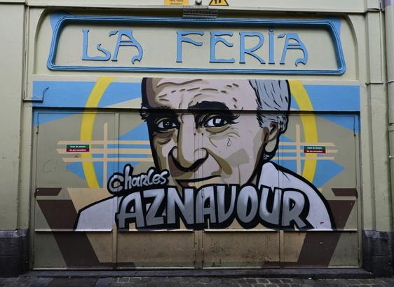 A mural (on a wall with a door) depicting a portrait of Charles Aznavour, with his name prominently displayed. The background features pastel colors and geometric shapes, and the sign above reads "LA FERIA."