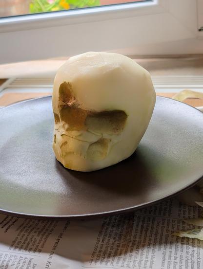Geschälter Kohlrabi auf einem dunklen Teller. Fraßspuren von Nacktschnecken lassen die Vorderseite wie einen menschlichen Schädel mit Augenhöhlen und Mund erscheinen. Im Hintergrund ein Fenster mit Tageslicht.