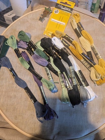 Photo of mocha coloured linen in a wooden hoop, placed across the top of the fabric are an assortment of embroidery threads, a pack of needles and a needle minder that is baby groot in his plant pot.