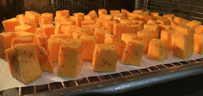 Cubes of sweet potato, oiled and seasoned with rosemary on baking paper in a tray in an oven, arranged in a tight grid.