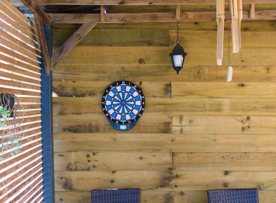 terrasse aménagée avec à gauche un claustra en lattes de bois et un pot de fleur suspendu, en face un mur de planches en clins avec une applique en forme de lanterne rustique et dessous une cible de fléchettes électronique. Plus bas on devine deux dossiers de fauteuil de jardin en plasti-rotin.