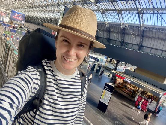 A person in a striped shirt and a straw hat smiles for a selfie while standing near a railing in a spacious train station. The background features a bustling scene with people, shops, and large screens displaying advertisements.