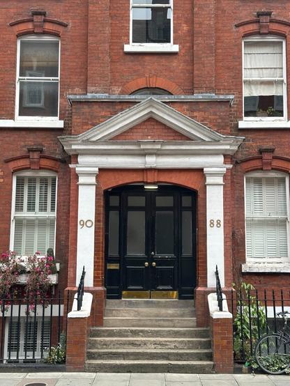 the main door on a brick multi-storey apartment building, possibly from the Victorian era. A stairway leads up to a portico elevated about half a storey above the ground, in which a black double door with a rounded top stands, framed by two neoclassical pilasters and topped by a triangular pediment, as if to divert attention from the somewhat less classical boxy brickwork behind it. The structure is surrounded by a constellation of sash windows, each under arched brickwork.