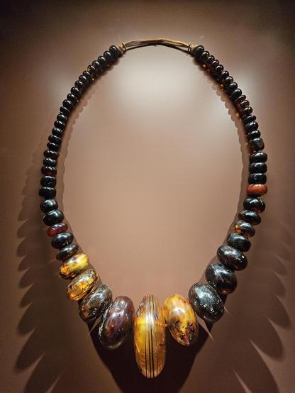 A large necklace made of polished amber beads in various shades from deep brown to golden yellow, strung in a gradient from small to large and back. The central beads are the largest and most vibrant, featuring natural inclusions and striations. The necklace hangs against a warm brown background.