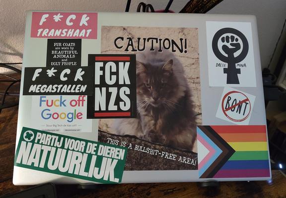Bestickerde laptop. Met de volgende stickers:
F*ck Transhaat, Fur coats are worn by beautyful animals and ugly people, F*ck Megastallen, Fuck off Google, Partij voor de Dieren Natuurlijk, FCK NZS, Caution this is a bullshit-free area met grumpy langharige kat, Dolle Mina, Bont verbodsbord, regenboogvlag.