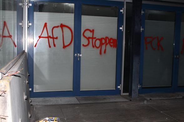 Rote Sprühfarbe bedeckt die Glasflächen mehrerer Türen. Die Worte 'AfD Stoppen' sind in großen, unregelmäßigen Buchstaben auf die mittleren Scheiben geschrieben. Links und rechts davon sind weitere Schriftzüge zu sehen. Die Türen sind von einem blauen Rahmen eingefasst, davor liegt ein Metallgitterboden. Im Vordergrund befindet sich ein Geländer, auf dem einige Kabel oder Drähte liegen. Unten links am Boden liegt ein kleiner, heller Gegenstand.