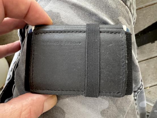 Ein kleines, schwarzes Portmonee, aus Papierleder und Stoff, verschlossen mit einem Gummiband, gehalten mit Zeigefinger und Daumen