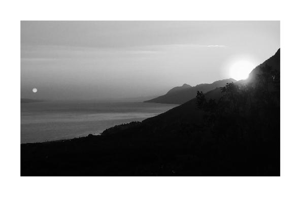 This black and white photo shows a stretch of coast of the Adriatic Sea in Croatia. It is late evening and the light is already very dim. In the right part of the picture the sun sets behind a mountain, in the left part of the photo you can see the rising moon. The sea with its horizon is visible.