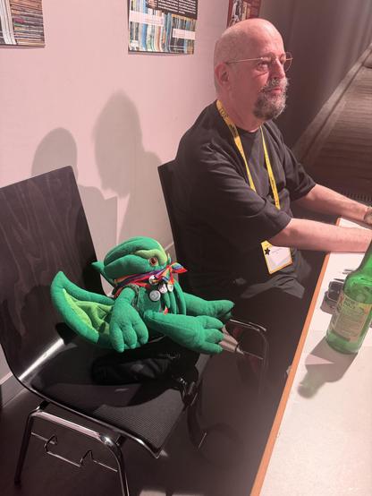 Fluff the Plush Cthulhu sitting at a table. Beyond Him is author Charles Stross