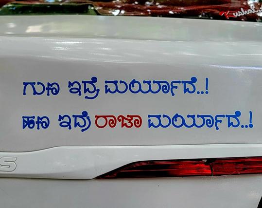 Close up of a blue bumper sticker on the back of a white car. The text is in Kannada:

ಗುಣ ಇದ್ರೆ ಮರ್ಯಾದೆ..!
ಹಣ ಇದ್ರೆ ರಾಜಾ ಮರ್ಯಾದೆ..!