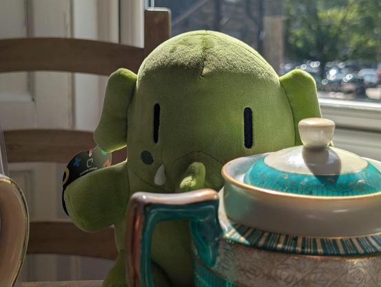 A green stuffed Mastodon elephant (about 20cm tall) peaking out from behind a teal tea pot.