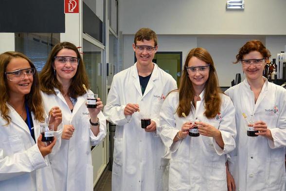 Fünf Schülerinnen und Schüler tragen Laborkittel und Schutzbrillen und führen in einem Labor einen chemischen Versuch mit Bechergläsern durch.