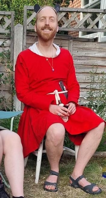 Photo of a man (me) sitting on a chair in a garden wearing a bright red tunic with a white shirt underneath, accessorised with a rope belt and a necklace.