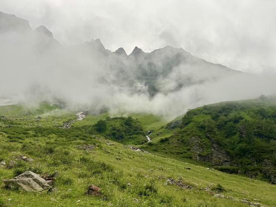 A misty mountainous landscape with lush green hills, rocky outcrops, and a snow meltwater stream flowing through the grassy terrain. Clouds partially obscure the mountain peaks in the background. Hohe Tauern.