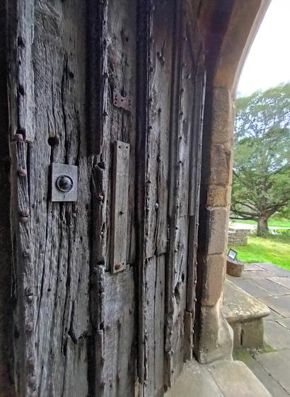 Photo of one half of large, double doorway. The door is very thick and heavy and is covered in large metal studs. A glimpse of a carved stone surround can also be seen, with a view of parkland beyond. The wood is ancient and weather worn with deep grooves. There are some amateur repairs where pieces of wood have been nailed on with no attempt to integrate them.