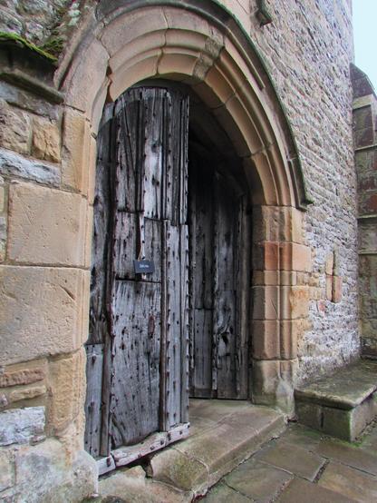 Photo of the double doored entrance to Haddon Castle and some of the courtyard wall. The doors are very thick and heavy and are covered in large metal studs. The left door, which includes a small portal is closed, the right door is open to visitors.
