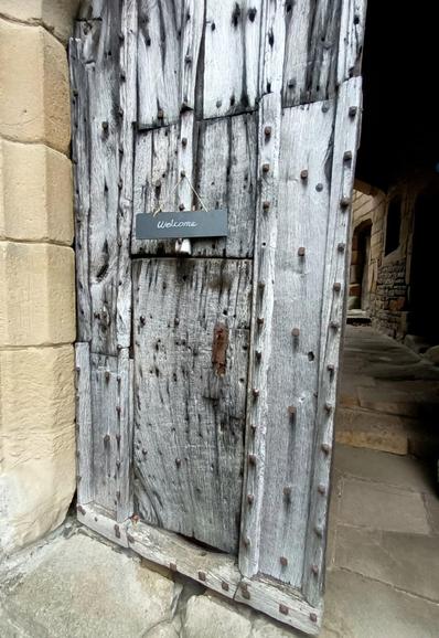 Photo of one half of a large, double doorway. The other door is open and out of shot but an old stone floor and tunnel into the couryard can be seen. The closed door is very thick and heavy and is covered in large metal studs and there is small door within the large door. A hand written sign above this says "Welcome" .