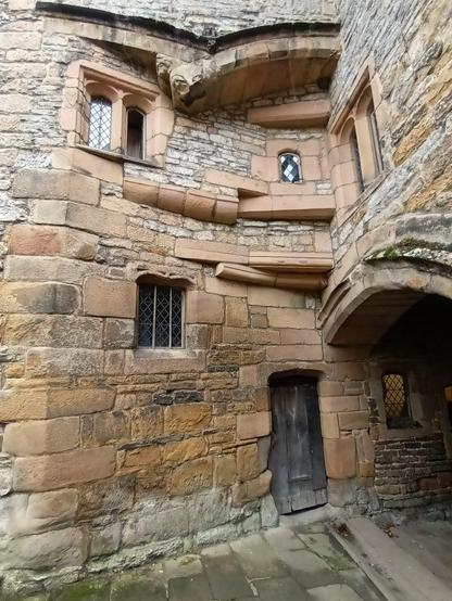 Photo showing a corner of a sandstone courtyard in a medieval castle. The architecture is intriguing. There is a small wonky door leading into a small room now used by the ticket inspector. Windows of all shapes and styles appear thrown together in a random, disorganized, or unplanned manner. The courses of stonework also give the same impression. To the right a low tunnel can be seen, this leads onto the main entrance shown in previous photos.