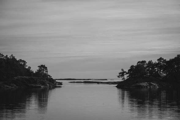 A greyscale monochrome photograph of strait between two small islands in an archipelago in the baltic sea. The stait is centered in the image. To the left and right on the edge of the image are rocky outcrops of two small islands behind them are two rows of small rocky islets. The water is calm with just some very small ripples.

Behind the last islet the open sea begins. 

An overcast, but quite light sky, with some horizontal structure in the clouds, dominates the upper half of the photograph.