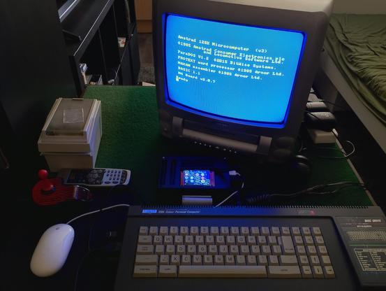 An Amstrad CPC6128 on a card table. An RSF3 expansion is connected to it, and a CRT TV displays the CPC boot screen with a series of optional ROM modules featuring a text editor, a machine code assembler and a module to enhance and support the expansion's USB ports.