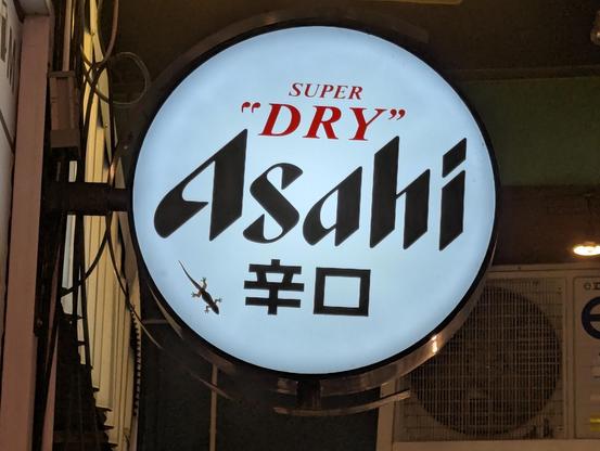 A sign advertising a drink named Asahi. It has a lizard standing on it, clinging to the surface. Lizard is "akesi" in toki pona.