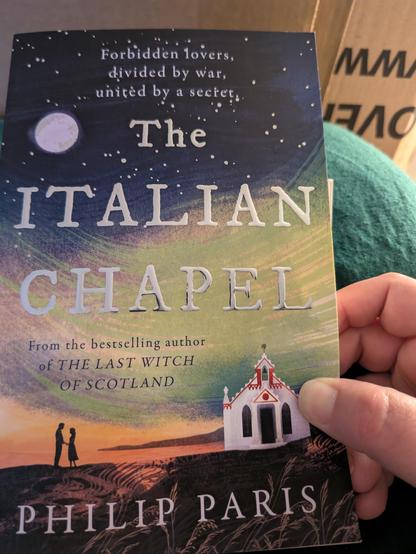 Front cover of the a book called The Italian Chapel by Philip Paris. The book shows the white chapel on dark ground, with the silhouette of two people standing close, the sky is shades of orange, green and blue with the moon visible.