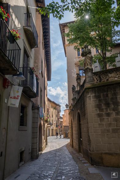 Color photo looking down a narrow cobblestone street in a Catalonian city center that is shaded in the foreground and sunlit beyond. To the right is a wall with statues of people on top of it and above them the is a rayed sunburst peeking through tree branches. In the distance are two people walking down the street.