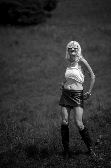 Blond woman hitching her black shiny skirt, making nice folds in the material. Her midrif is bared by her white top, Her shoulders and arms show sinews and muscles under mature skin. She stads in a meadow that climbs a hill behind her. The grey  shades are lovely.