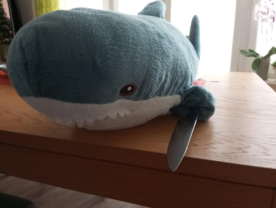 Blåhaj on a table facing the camera with a knife in fin.

Credits: u/TheSimpofDarkness