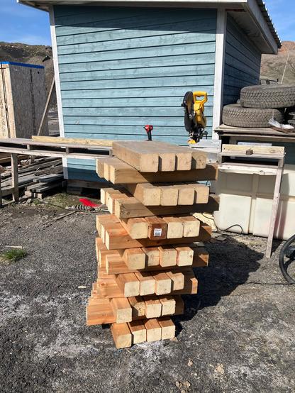 A stack of short 4x4s, four in each of the thirteen layers. Behind the stack there is a mitre saw on a stand and a blue shed.