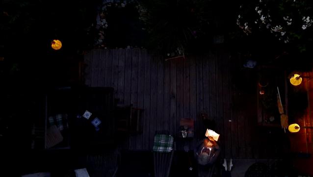 Looking at my porch from above. Some clutter is unvisible in the dark. Three lanterns give a warm yellowish light, a person sits reading in the dark. One easy xhair has just been vacated, a checken cushion refelts a bit of light.